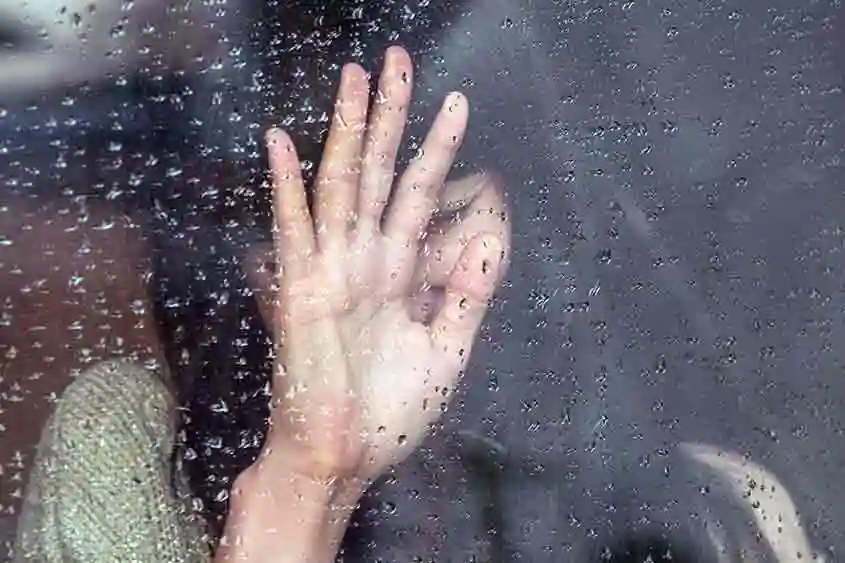 Image of a woman with closed eyes, and a hand raised to a window with water droplets on the glass surface. It could possibly imply depression